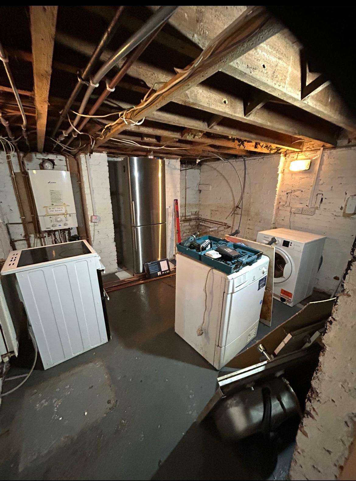 Old basement utility room with exposed pipes and scattered appliances before renovation